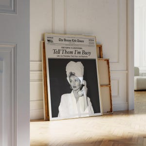 May include: A framed black and white print featuring a woman wearing a towel on her head and holding a telephone. The print has the headline "Tell Them I'm Busy" and the newspaper title "The Dream Life Times". The print is displayed in a room with a wooden floor.