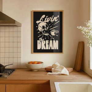 May include: A framed black and white art print with the words "Livin' the Dream" and a skull holding a coffee cup. The print is in a wooden frame and hangs on a beige wall above a wooden countertop. A bowl of oranges and a cutting board are on the counter.