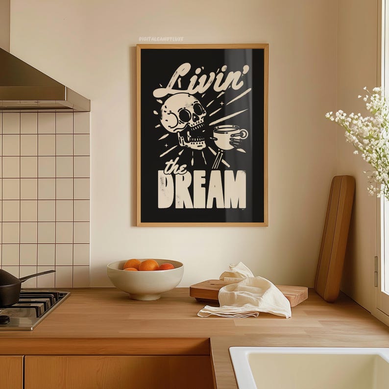 May include: A framed black and white art print with the words "Livin' the Dream" and a skull holding a coffee cup. The print is in a wooden frame and hangs on a beige wall above a wooden countertop. A bowl of oranges and a cutting board are on the counter.
