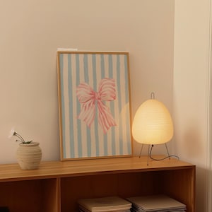 May include: A framed print of a pink bow on a blue and white striped background. The print is hanging on a wall above a wooden shelf with a white lamp on the shelf.