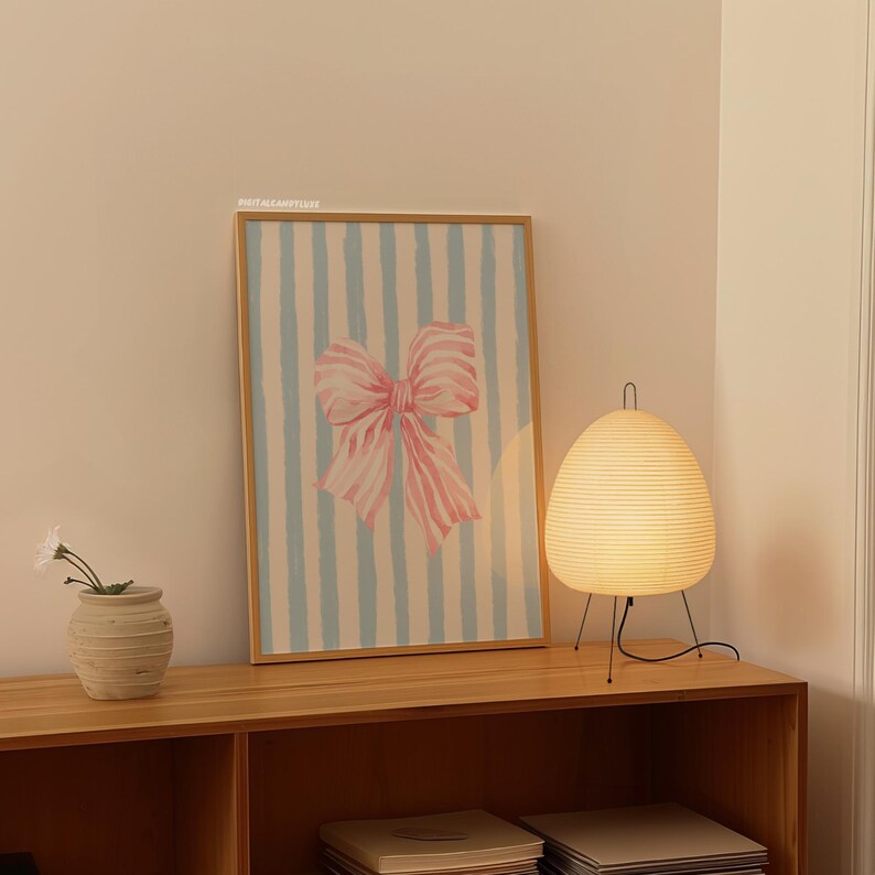May include: A framed print of a pink bow on a blue and white striped background. The print is hanging on a wall above a wooden shelf with a white lamp on the shelf.