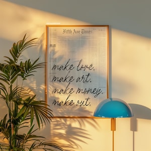 May include: A framed print with the text "Fifth Ave Times." and the words "make love. make art. make money. make out." in a handwritten style. The frame is a light brown color, and the background is a newspaper print.