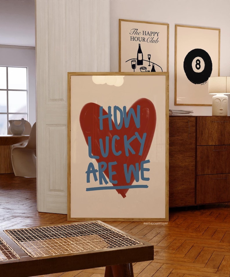 May include: A framed art print with a red heart and the words "HOW LUCKY ARE WE" in blue. Two other framed prints are visible, one with a black eight ball and another with the text "The Happy Hour Club". The room has wood floors and a wooden cabinet.