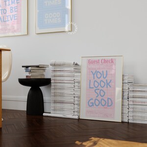 May include: A framed print with the text "You look so good" on a pink and white guest check. The print is framed in gold and is leaning against a stack of magazines.