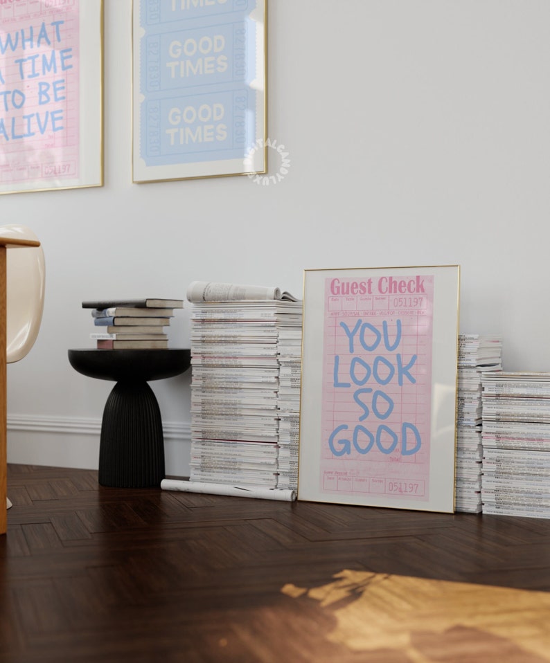 May include: A framed print with the text "You look so good" on a pink and white guest check. The print is framed in gold and is leaning against a stack of magazines.