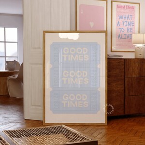 May include: A framed print with a light blue background and white text that reads "Good Times" repeated three times. The print is in a wooden frame and is hanging on a wall in a room with a wooden floor.