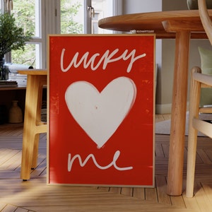 May include: A framed red canvas print with white lettering that says "lucky me" and a white heart shape in the center.