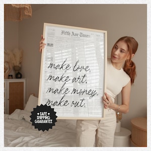 May include: A framed print with the text "make love, make art, make money, make out" in a handwritten style. The background resembles a newspaper. The frame is light-colored. The print is held by a person.