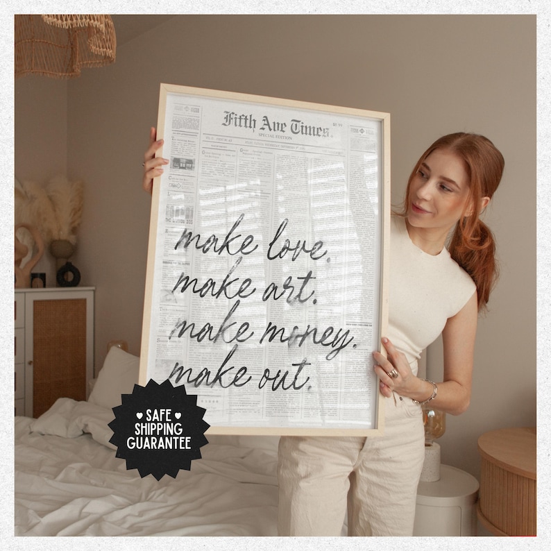 May include: A framed print with the text "make love, make art, make money, make out" in a handwritten style. The background resembles a newspaper. The frame is light-colored. The print is held by a person.