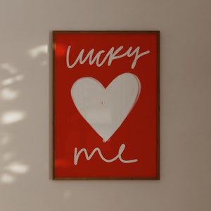 May include: A framed print with a red background and white text that says "lucky me". A white heart is drawn in the center of the print.