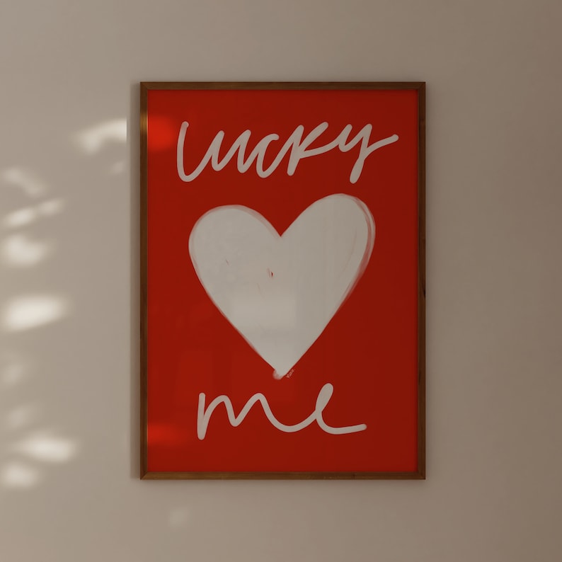 May include: A framed print with a red background and white text that says "lucky me". A white heart is drawn in the center of the print.