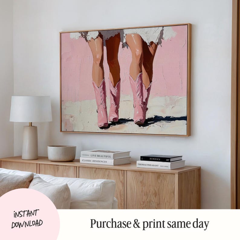 May include: A painting of two pairs of legs wearing pink cowboy boots against a pink background. The painting is framed in a light wood frame and hanging on a white wall.
