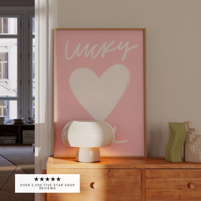 May include: A white ceramic mushroom lamp with a pink and white framed print that says "lucky" with a white heart in the center. The print is on a wooden dresser with two drawers.