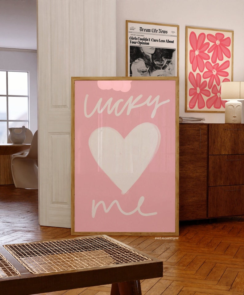 May include: Pink and white art print with a heart and the words "lucky me" in white script. The print is framed in a light wood frame and is hanging on a wall.