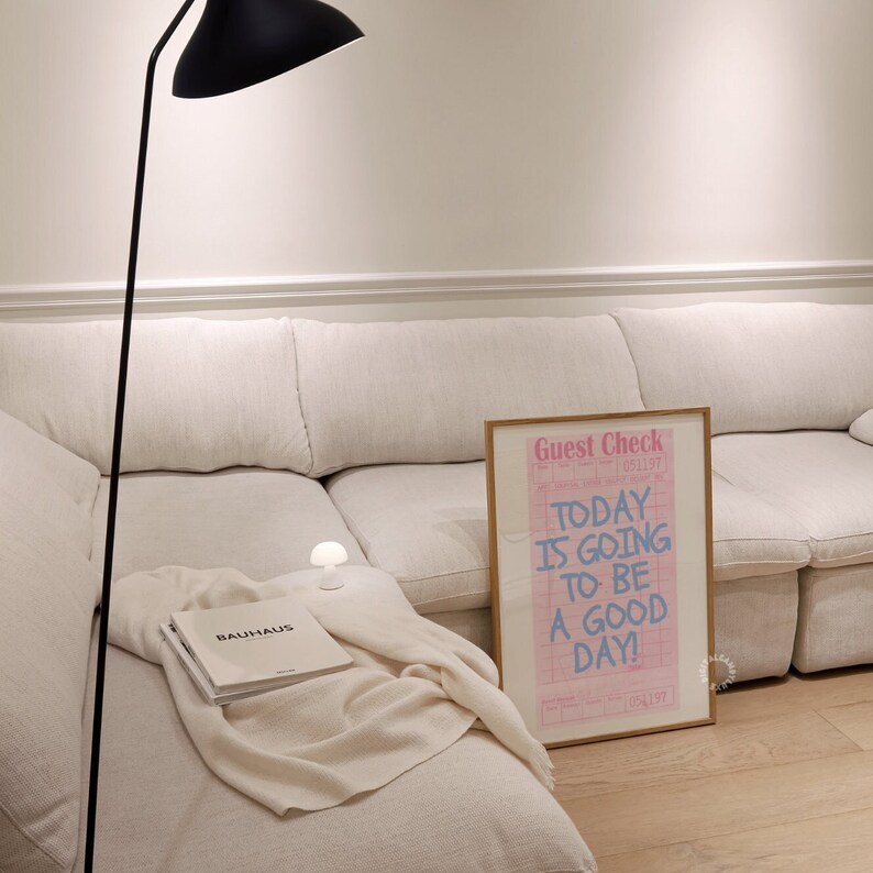 May include: A white sectional sofa with a throw blanket and a book on it. A framed poster with the text "Today is going to be a good day!" is leaning against the sofa.