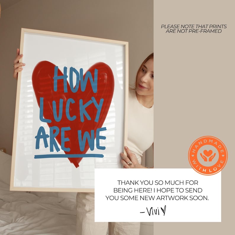 May include: Framed artwork featuring a large red heart with the words "HOW LUCKY ARE WE" in blue. The artwork is held by a person. The frame is light-colored. The image also includes a small orange "HANDMADE WITH LOVE" sticker.