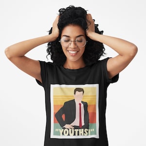 Könnte beinhalten: Schwarzes T-Shirt mit einer Grafik eines Mannes im Anzug und roter Krawatte vor einem Retro-Sonnenuntergang. Der Text "YOUTHS!" steht unter der Grafik. Die Person, die das T-Shirt trägt, lächelt.