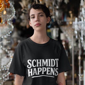 Può includere: T-shirt nero con la scritta bianca "SCHMIDT HAPPENS". La persona indossa la maglietta. Lo sfondo è un negozio con lampadari e altri apparecchi di illuminazione.