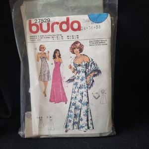 May include: A vintage Burda sewing pattern, size 34+36+38, featuring illustrations of three different dress designs. The pattern includes a long, floral print dress, a pink dress, and a floral print dress with a shawl. The pattern is in a plastic sleeve.