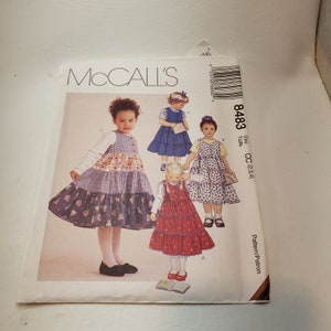 May include: McCall's sewing pattern for children's dresses. The pattern features illustrations of four different tiered dresses in various colors and patterns. The pattern number is 8483, and the size is CC (2,3,4).