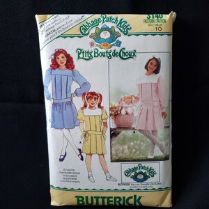 May include: A sewing pattern for Cabbage Patch Kids clothing. The pattern includes instructions for making two dresses, one blue and one pink, with a white collar and a bow. The pattern is size 10 and includes a bonus iron-on transfer.