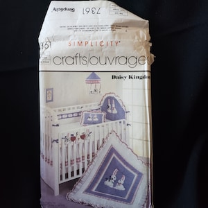 Op de afbeelding: Een Simplicity crafts/ouvrages-patroon voor een babybed-beddengoedset met een Daisy Kingdom-thema. Het patroon omvat een bedrok, een deken, een kussen en een hemel. De deken heeft een blauwe achtergrond met witte konijnen en een witte rand.