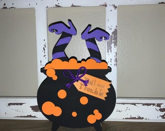May include: A black cauldron with orange accents and witch legs. The cauldron has orange bubbles and a tag that reads "Toil and Trouble". The witch legs are purple with black stripes. A Halloween-themed decoration.