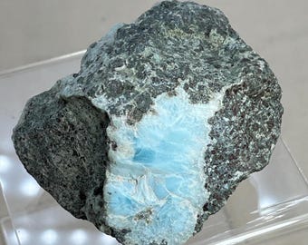 May include: A raw mineral specimen featuring a rough, dark grey, textured exterior matrix surrounding a central, smooth, sky-blue crystalline inclusion with visible white veining and a glossy, polished appearance on the exposed surface.