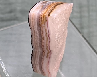 May include: A polished mineral specimen featuring vertical bands of soft pink, white, and earthy brown tones. The side displays a rough, crystalline texture with a jagged edge, while the front face is smooth and flat, showcasing natural layers.