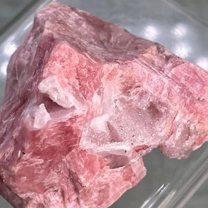 May include: A rough-cut, natural rhodochrosite specimen. The stone displays a vibrant pink hue with areas of white quartz. The crystalline structure is visible, showcasing the mineral's natural formation. The rhodochrosite is presented in a clear, rectangular display case.