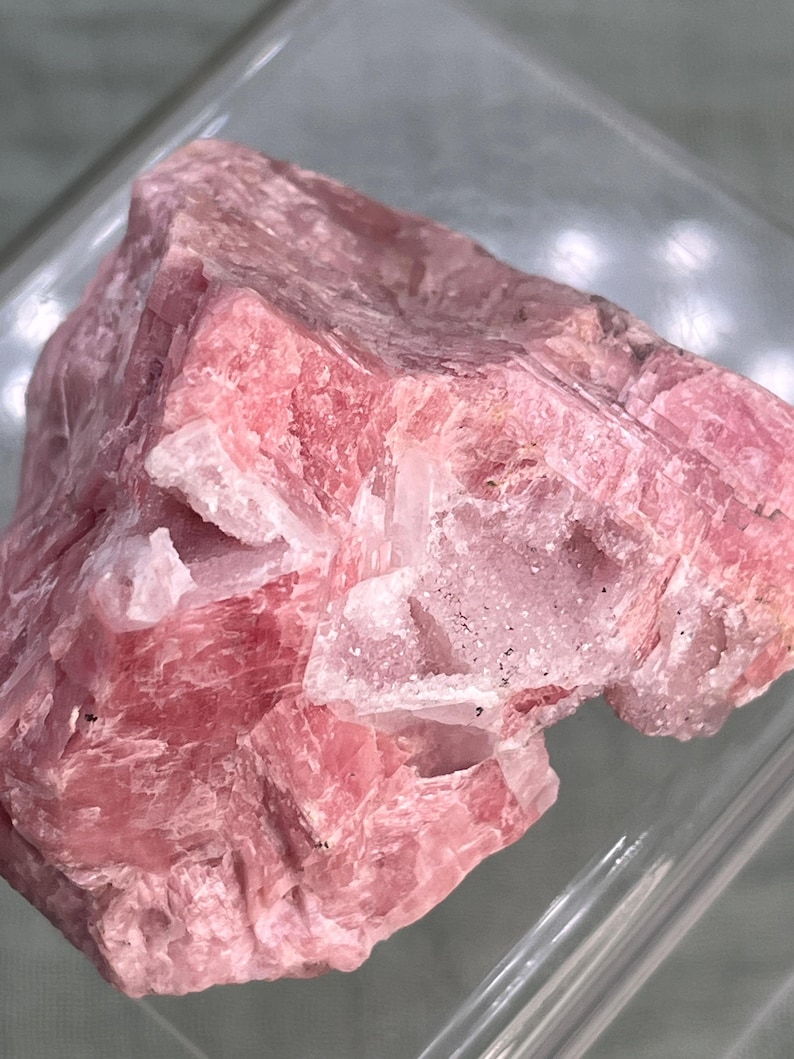 May include: A rough-cut, natural rhodochrosite specimen. The stone displays a vibrant pink hue with areas of white quartz. The crystalline structure is visible, showcasing the mineral's natural formation. The rhodochrosite is presented in a clear, rectangular display case.