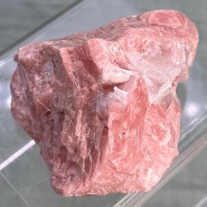 May include: A raw, uncut piece of rhodochrosite, a pink manganese carbonate mineral. The stone has a layered, crystalline structure with varying shades of pink and white. The rough texture and natural form of the stone are visible.