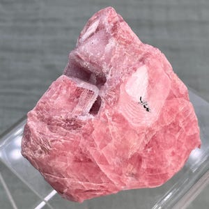 May include: A raw, uncut rhodochrosite gemstone. The stone is a vibrant pink color with a crystalline structure and a rough, textured surface. The gemstone has a natural, organic shape and is displayed on a clear, angled surface.