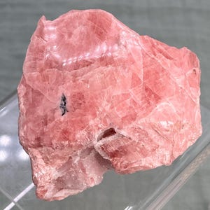 May include: A rough, pink rhodochrosite mineral specimen. The stone has a layered appearance with varying shades of pink and some darker inclusions. The surface is textured, and the overall shape is irregular.