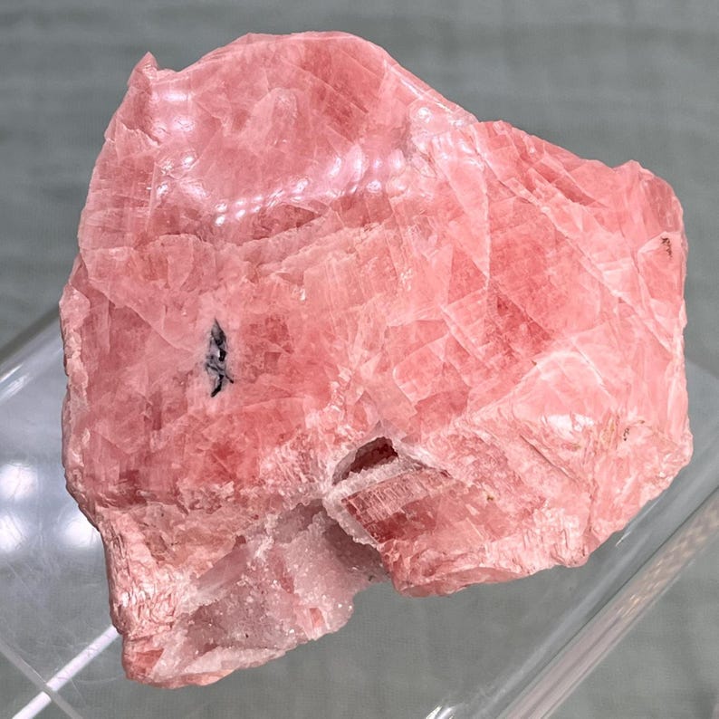 May include: A rough, pink rhodochrosite mineral specimen. The stone has a layered appearance with varying shades of pink and some darker inclusions. The surface is textured, and the overall shape is irregular.