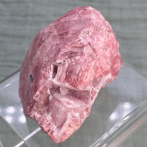 May include: A rough, natural rhodochrosite gemstone. The stone is a vibrant pink color with white and clear crystalline inclusions. The surface has a textured, layered appearance. The gemstone is displayed on a clear, rectangular stand.