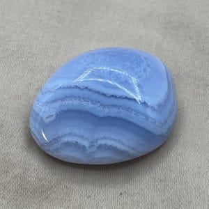May include: A smooth, oval-shaped blue lace agate gemstone. The stone features bands of light and dark blue, creating a layered, wave-like pattern. The polished surface reflects light, highlighting the stone's natural beauty.