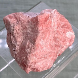 May include: A rough, raw piece of rhodochrosite, a pink mineral. The stone has a layered, textured appearance with varying shades of pink. The mineral is displayed on a clear, angled surface.