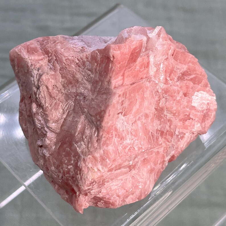 May include: A rough, raw piece of rhodochrosite, a pink mineral. The stone has a layered, textured appearance with varying shades of pink. The mineral is displayed on a clear, angled surface.