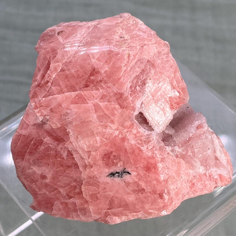 May include: A rough-cut, pink mineral specimen with a crystalline structure. The stone has a layered appearance with variations in color, ranging from light to deep pink. A small, dark inclusion is visible on the surface. The mineral is displayed on a clear, angled surface.