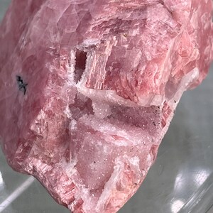 May include: Close-up of a pink mineral specimen. The stone has a layered, crystalline structure with varying shades of pink and some white inclusions. The surface is rough and textured, with visible lines and patterns.