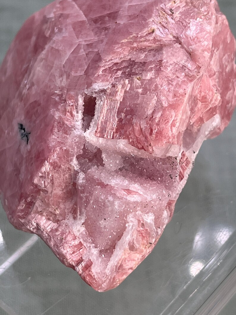 May include: Close-up of a pink mineral specimen. The stone has a layered, crystalline structure with varying shades of pink and some white inclusions. The surface is rough and textured, with visible lines and patterns.