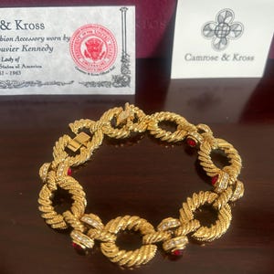 May include: A gold-tone link bracelet featuring a twisted rope texture, accented with small clear crystals and round red stones. The bracelet is displayed with two white cards, one featuring the text Camrose & Kross and a seal of the President of the United States.