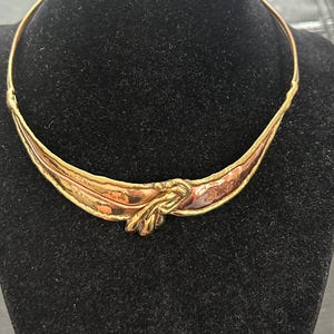 May include: A rigid gold-tone metal collar necklace featuring a central knot detail. The design incorporates a mix of polished gold-tone and textured copper-tone metal sections that curve along the front, creating a layered, organic appearance.