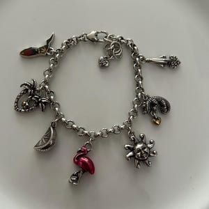May include: A silver charm bracelet with various tropical-themed charms. Charms include a flamingo, sun, palm tree, slice of citrus, and a shoe. The bracelet is on a white surface.