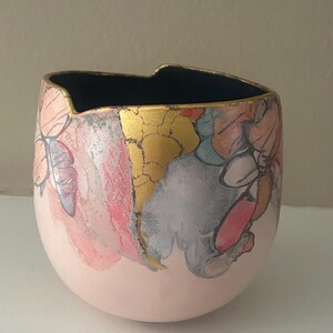 May include: A hand-painted ceramic bowl with a rounded shape and a wide opening. The exterior features a light pink base with colorful abstract designs in pink, gold, and gray. The interior is black, and the rim is accented with gold.