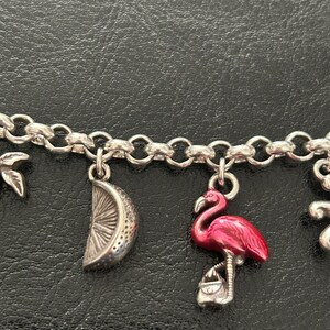 May include: Silver charm bracelet with a flamingo charm in bright pink enamel, a silver lemon slice charm, and other charms. The bracelet is on a black surface.