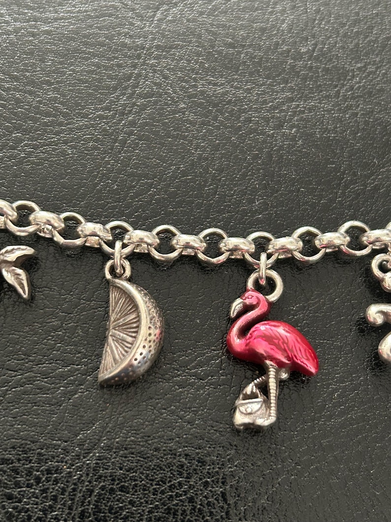 May include: Silver charm bracelet with a flamingo charm in bright pink enamel, a silver lemon slice charm, and other charms. The bracelet is on a black surface.