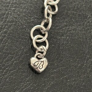 May include: Silver-toned necklace with a heart-shaped charm. The charm features the letter "B" in a cursive script. The necklace is composed of interconnected circular links. The background is a dark, textured surface.