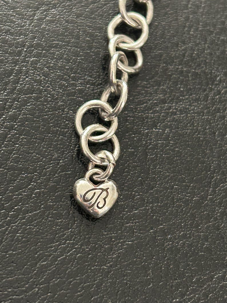 May include: Silver-toned necklace with a heart-shaped charm. The charm features the letter "B" in a cursive script. The necklace is composed of interconnected circular links. The background is a dark, textured surface.